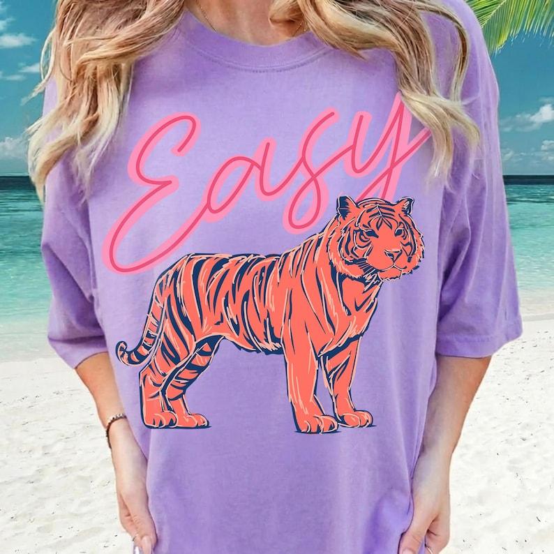 May include: A lilac-colored t-shirt featuring a vibrant orange tiger illustration with navy stripes. Above the tiger, the word "Easy" is written in a cursive, neon pink font. The shirt has short sleeves and a relaxed fit.