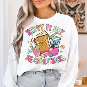 May include: White long-sleeve sweatshirt with a colorful graphic design that says "Happy Pi Day Be Irrational". The graphic includes a notebook, pencils, a calculator, a ruler, and an apple.