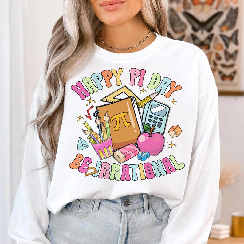 May include: White long-sleeve sweatshirt with a colorful graphic design that says "Happy Pi Day Be Irrational". The graphic includes a notebook, pencils, a calculator, a ruler, and an apple.