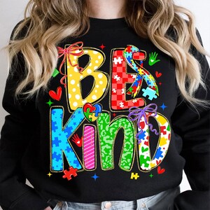 May include: Black sweatshirt with the words "BE KIND" in colorful, patterned letters. The design includes puzzle pieces, hearts, and bows, with a focus on autism awareness. The sweatshirt is a crew neck style.