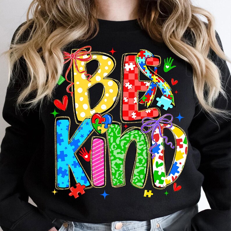 May include: Black sweatshirt with the words "BE KIND" in colorful, patterned letters. The design includes puzzle pieces, hearts, and bows, with a focus on autism awareness. The sweatshirt is a crew neck style.