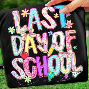 May include: Black t-shirt with the text "LAST DAY OF SCHOOL" in colorful, whimsical lettering. The design includes flowers, a pencil, an apple, a globe, a paper airplane, and bows, creating a festive back-to-school theme.