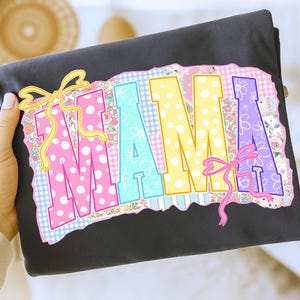May include: A black t-shirt featuring the word "MAMA" in large, colorful letters. The letters are decorated with polka dots, floral patterns, and bows. The design has a patchwork style with a mix of pastel colors.