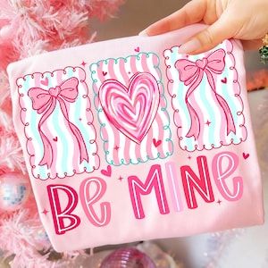 May include: A light pink t-shirt with a Valentine's Day design. The design includes three framed images with a heart and bows, and the words "Be Mine" in pink and red lettering.