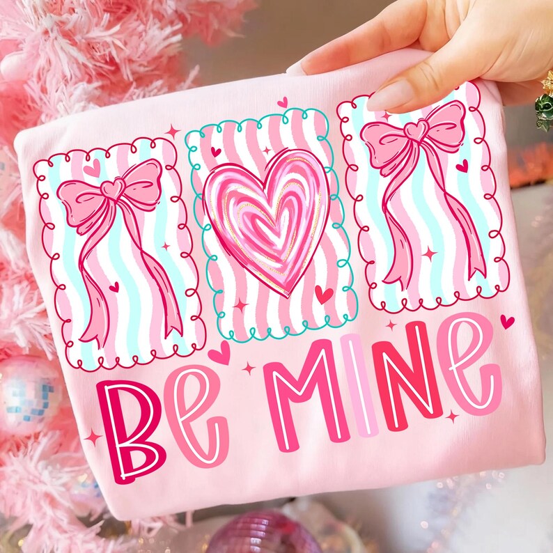 May include: A light pink t-shirt with a Valentine's Day design. The design includes three framed images with a heart and bows, and the words "Be Mine" in pink and red lettering.