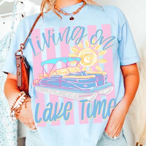 May include: Light blue t-shirt with a graphic of a pontoon boat, sun, and text "Living on Lake Time." The design features a blue boat with a yellow canopy and a yellow sun on a pink and blue striped background. The shirt is a casual wear item.