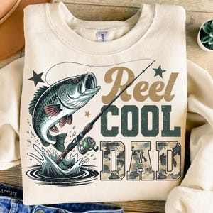 Puede incluir: Sudadera de color crema con un diseño gráfico que presenta un pez saltando del agua, una caña de pescar y el texto "Reel Cool Dad" en marrón y verde. La palabra "DAD" tiene un patrón de camuflaje.