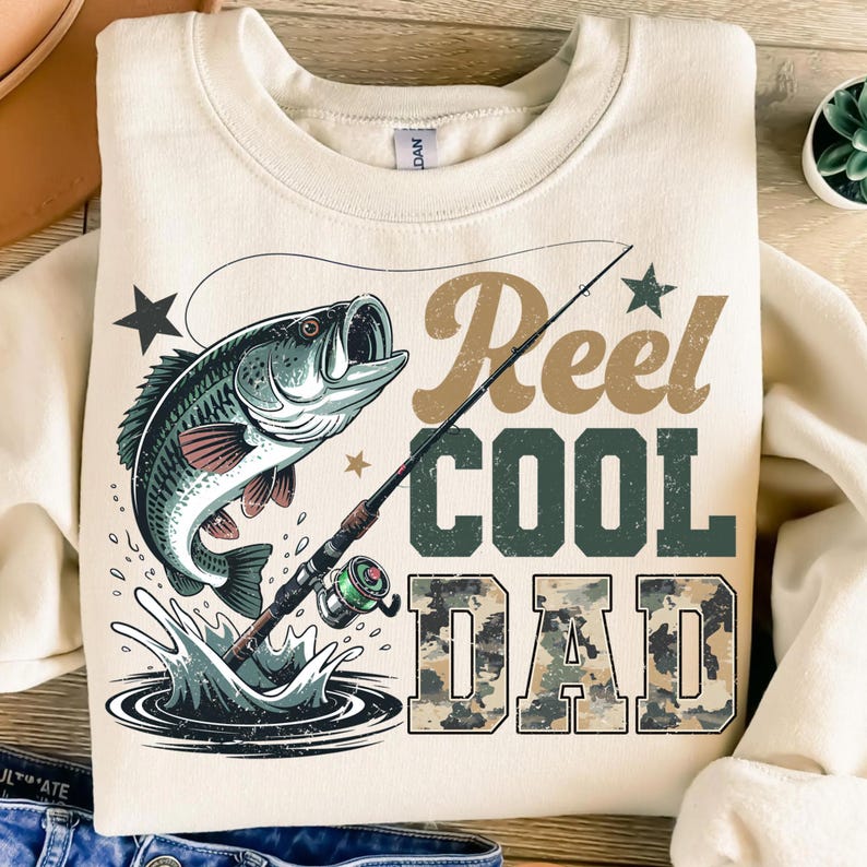 Może przedstawiać: Kremowa bluza z nadrukiem przedstawiającym rybę wyskakującą z wody, wędkę oraz napis "Reel Cool Dad" w kolorze brązowym i zielonym. Słowo "DAD" ma wz&oacute;r kamuflażu.