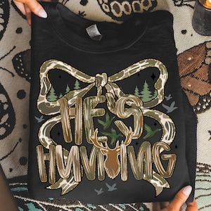 May include: Black sweatshirt with a camouflage bow design and the text "HE'S HUNTING". The graphic includes a deer head, trees, and flying birds. The sweatshirt is held by two hands.
