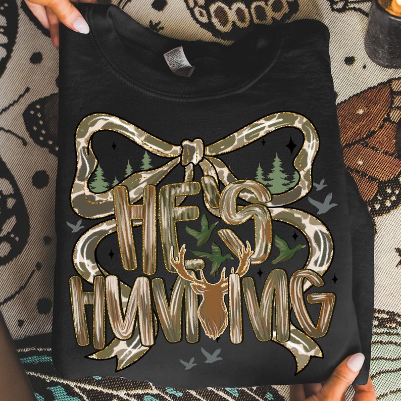 May include: Black sweatshirt with a camouflage bow design and the text "HE'S HUNTING". The graphic includes a deer head, trees, and flying birds. The sweatshirt is held by two hands.