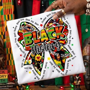 May include: White t-shirt featuring a colorful bow design with the words "BLACK History" in bold letters. The bow incorporates images of Black women, flowers, and geometric patterns. The words "Black Love," "Black Joy," and "Black Pride" are also included.