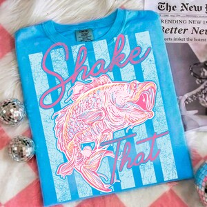 K&ouml;nnte beinhalten: T&uuml;rkisfarbenes Langarm-T-Shirt mit Used-Design. Das Shirt zeigt eine rosa-orangefarbene Fischgrafik mit den Worten "Shake That" in rosa Schrift. Hinter dem Fisch befinden sich vertikale hellblaue Streifen.