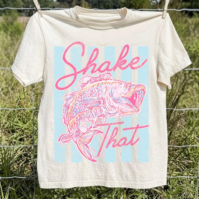 K&ouml;nnte beinhalten: Cremefarbenes T-Shirt mit einer rosa-blauen Grafik eines Fisches und den Worten "Shake That". Der Fisch ist mit rosa, orange und gelben Akzenten versehen, vor einem hellblau gestreiften Hintergrund.
