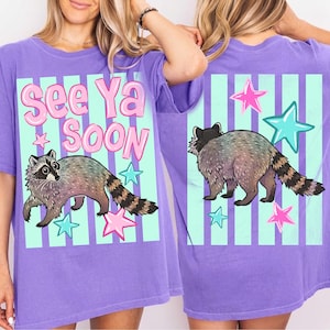 Puede incluir: Camiseta morada con un diseño de mapache y el texto "See Ya Soon". El diseño incluye estrellas rosas y turquesas y rayas verticales. El mapache tiene un cuerpo gris y marrón con una cola a rayas.
