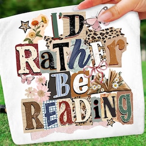 May include: White decorative pillow with the text "I'd Rather Be Reading" in a collage style. The design includes floral and butterfly accents, and a star-shaped embellishment. The pillow is held by a hand.