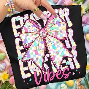 May include: Black t-shirt with a large, colorful bow design featuring pastel Easter eggs, bunnies, and flowers. The words "Easter Vibes" are in pink script. The design has a checkered pattern.