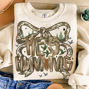 May include: Cream-colored sweatshirt with the text "HE'S HUNTING" in a decorative font, featuring a deer head and a bow design with a camouflage pattern. The design includes trees and birds.