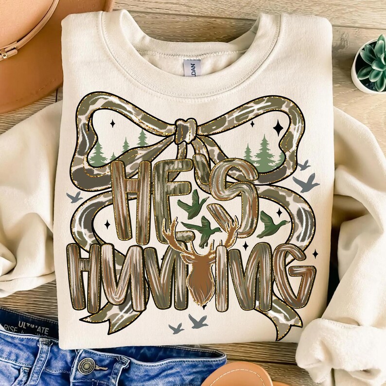 May include: Cream-colored sweatshirt with the text "HE'S HUNTING" in a decorative font, featuring a deer head and a bow design with a camouflage pattern. The design includes trees and birds.