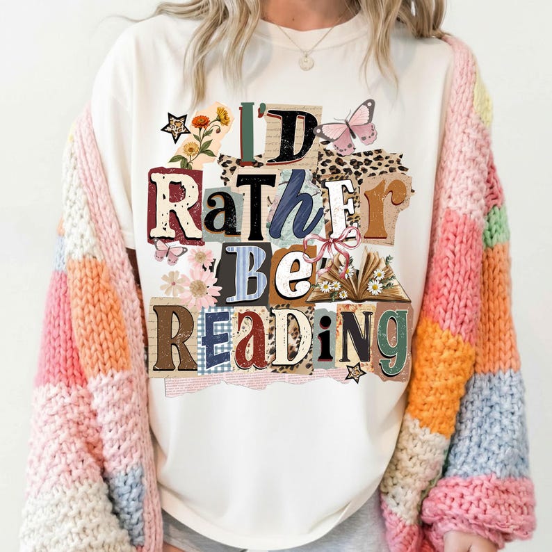 May include: Cream-colored t-shirt with the text "I'D RATHER BE READING" in a collage style with floral and butterfly accents. The person is wearing a colorful striped cardigan.