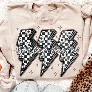 Puede incluir: Sudadera beige con un dise&ntilde;o gr&aacute;fico que presenta tres rayos con balones de f&uacute;tbol y un patr&oacute;n de cuadros blancos y negros. El texto "Soccer Mom" est&aacute; escrito sobre los rayos.