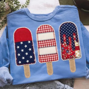 May include: Pale blue sweatshirt with three appliqu&eacute;d popsicle designs. The popsicles feature star and stripe, gingham, and floral patterns. The popsicle sticks are made of burlap. The design is reminiscent of the American flag.