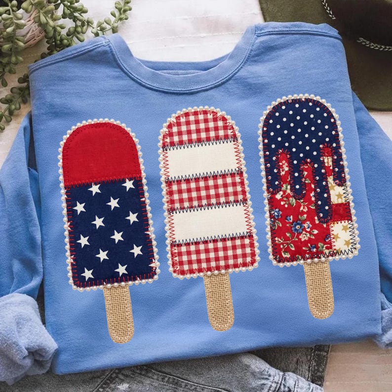 May include: Pale blue sweatshirt with three appliqu&eacute;d popsicle designs. The popsicles feature star and stripe, gingham, and floral patterns. The popsicle sticks are made of burlap. The design is reminiscent of the American flag.