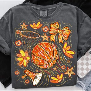 May include: Dark gray t-shirt with a basketball-themed graphic. The design includes an orange basketball, a basketball hoop, flowers, stars, a bow, and a megaphone. The graphic has orange, yellow, and gold accents.