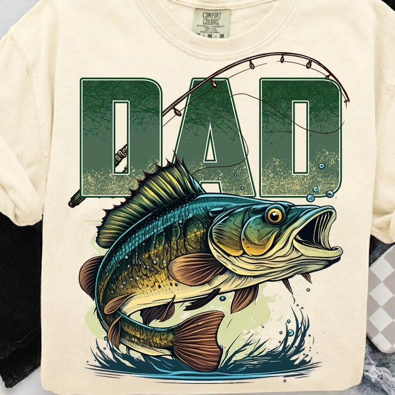Pu&ograve; includere: T-shirt color crema con la scritta "DAD" in verde e una canna da pesca. Sotto, un'illustrazione dettagliata di un pesce con la bocca aperta, spruzzi d'acqua e toni marroni e blu.