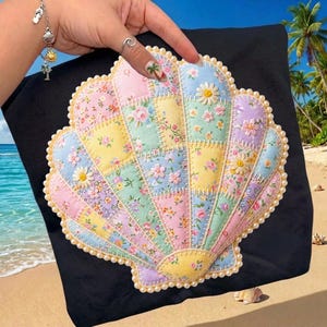 Puede incluir: Un diseño de concha de patchwork colorido sobre un fondo negro. La concha está hecha de cuadrados de tela de colores pastel con estampados florales y acentos de margaritas. Los bordes están adornados con un borde de perlas.