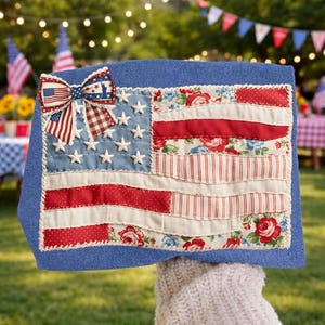 May include: A blue decorative pillow featuring a patchwork American flag design. The flag includes red and white stripes, a blue field with white stars, and a bow with an American flag pattern. Floral and striped fabric accents add detail.