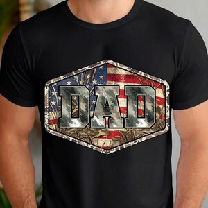 以下が含まれることがあります： 黒いTシャツ。六角形のデザインで、迷彩柄の「DAD」の文字が描かれています。デザインにはアメリカ国旗と迷彩柄が組み込まれており、愛国心とアウトドアをテーマにしています。