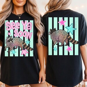 May include: Black t-shirt featuring a raccoon illustration with the text "See Ya Soon". The design includes pink and blue stars and is set against a light green striped background. The raccoon has a striped tail.