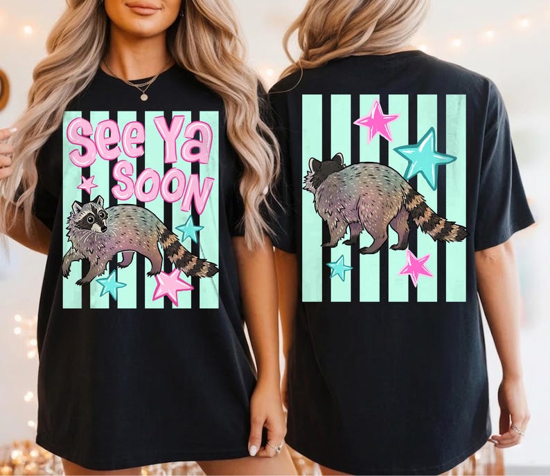 May include: Black t-shirt featuring a raccoon illustration with the text "See Ya Soon". The design includes pink and blue stars and is set against a light green striped background. The raccoon has a striped tail.
