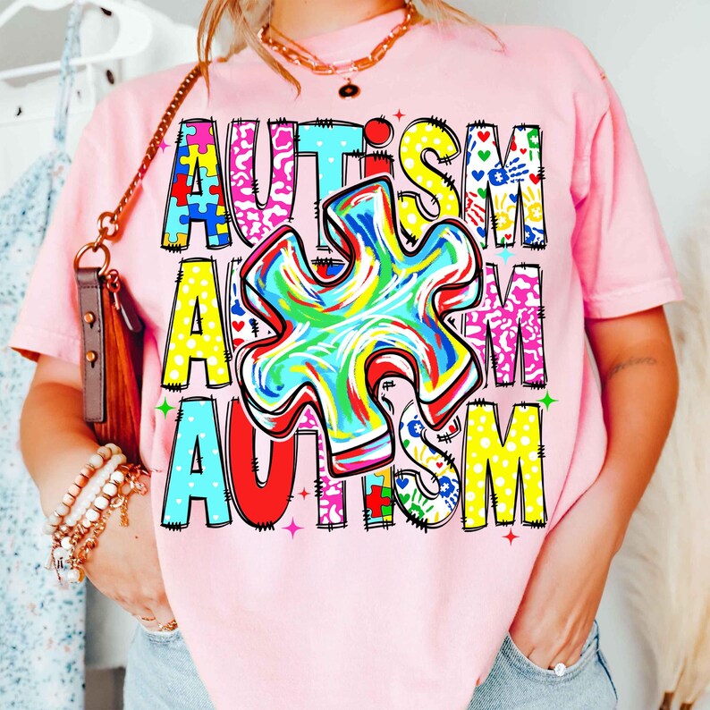 May include: Pink t-shirt with the word "AUTISM" in colorful, block letters. The "U" in the word is a multi-colored puzzle piece. The shirt has a crew neck and short sleeves.