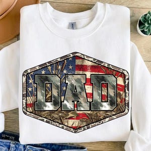 以下が含まれることがあります： 「DAD」の文字が大きくプリントされた白いスウェットシャツ。グラフィックは迷彩柄で、背景にはアメリカ国旗が描かれています。柔らかい素材で作られたスウェットシャツです。