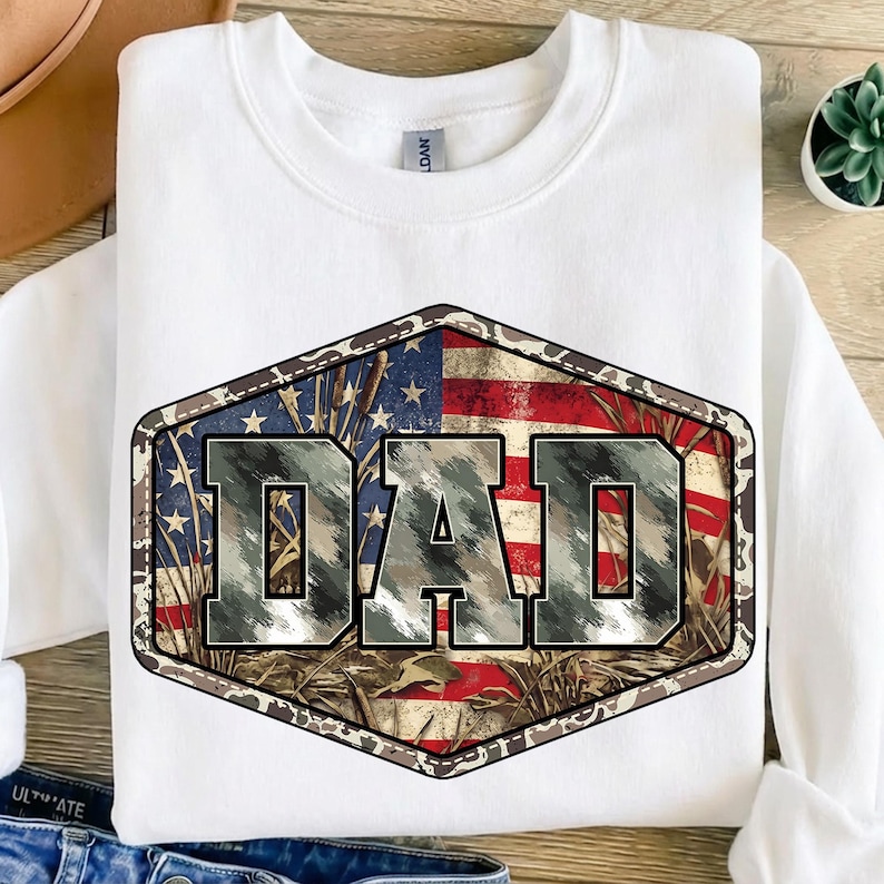 以下が含まれることがあります： 「DAD」の文字が大きくプリントされた白いスウェットシャツ。グラフィックは迷彩柄で、背景にはアメリカ国旗が描かれています。柔らかい素材で作られたスウェットシャツです。