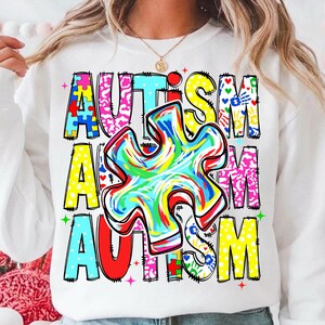 May include: White sweatshirt with the word "AUTISM" in colorful, block letters. A large, multi-colored puzzle piece is centered on the front. The design includes various patterns and handprints.