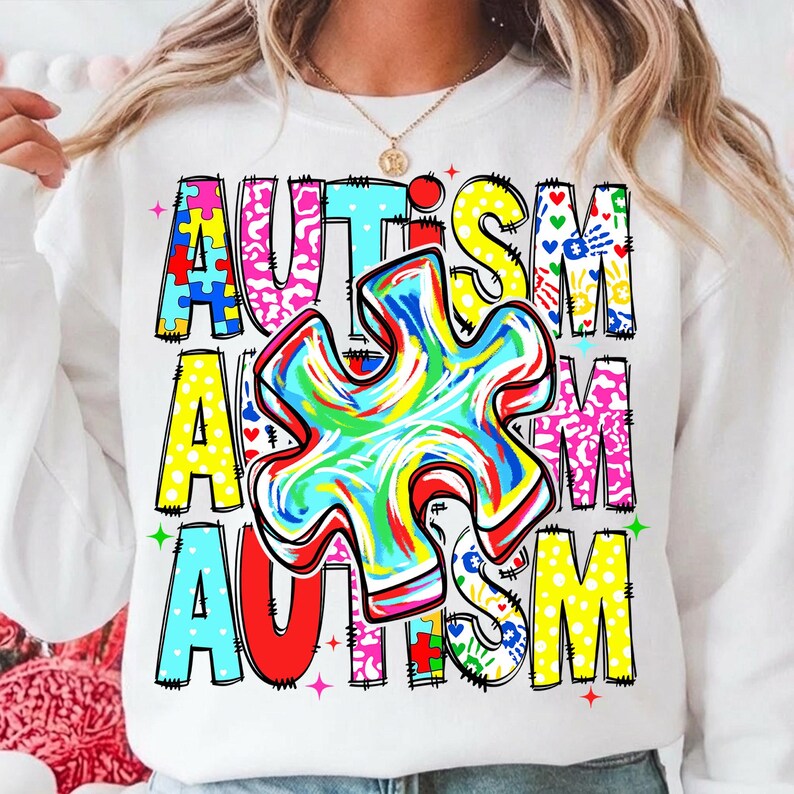 May include: White sweatshirt with the word "AUTISM" in colorful, block letters. A large, multi-colored puzzle piece is centered on the front. The design includes various patterns and handprints.