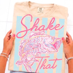 K&ouml;nnte beinhalten: Beiges T-Shirt mit rosa und blauem Grafikdesign. Das Design zeigt eine gro&szlig;e Fischillustration mit den Worten "Shake That" in rosa Schrift. Das Shirt hat einen Rundhalsausschnitt und ein Etikett oben in der Mitte.
