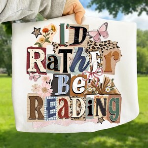 May include: White tote bag with the text "I'd Rather Be Reading" in a collage style. The design includes butterflies, flowers, and an open book. The letters are in various colors and fonts.