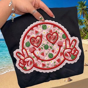 Può includere: T-shirt nero con un design a faccina sorridente a tema anguria. Il design include fette di anguria, cuori e segni di pace, tutti bordati di rosso e bianco. La faccina sorridente ha occhi a forma di cuore e una bocca sorridente.