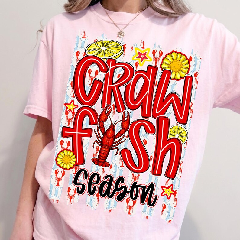 May include: A light pink t-shirt with a graphic that says "Crawfish Season" in red, outlined in white and black. The design includes a red crawfish, yellow lemons, stars, and sunflowers. The background has a pattern of red crawfish on a light blue and white striped background.