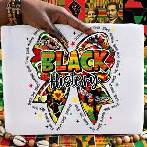 May include: White t-shirt featuring a colorful bow design with the words "BLACK History" in bold letters. The bow incorporates various patterns and images, with additional text around the bow. The shirt is held by a hand.