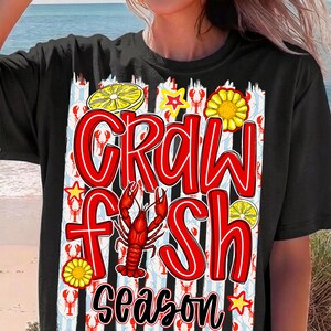 May include: Black t-shirt with a colorful graphic that reads "Crawfish Season." The design includes a red crawfish, lemons, and star shapes. The background has a pattern of red crawfish and white and blue stripes.