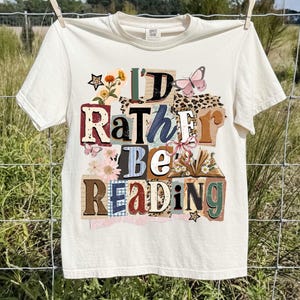 May include: Cream-colored t-shirt with the text "I'd Rather Be Reading" in a collage of colorful letters, flowers, and butterflies. The shirt is hanging on a clothesline.