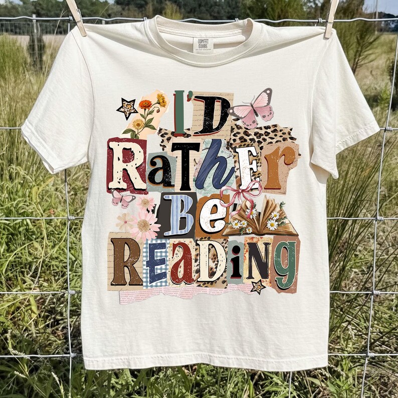 May include: Cream-colored t-shirt with the text "I'd Rather Be Reading" in a collage of colorful letters, flowers, and butterflies. The shirt is hanging on a clothesline.
