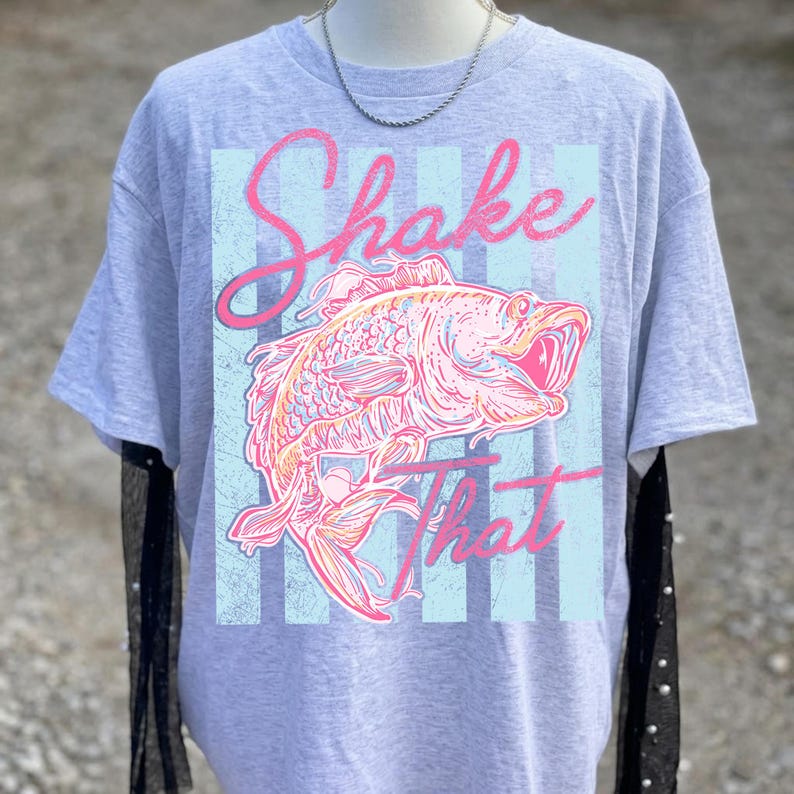 K&ouml;nnte beinhalten: Graues T-Shirt mit einem rosa-orangenen Fisch-Design und dem pinkfarbenen Schriftzug "Shake That". Der Fisch ist vor einem hellblauen Hintergrund mit vertikalen Streifen platziert.