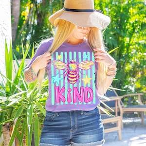 Puede incluir: Una camiseta morada con un gr&aacute;fico de abeja colorida, rayas turquesas y blancas, y la palabra "KIND" en rosa. La abeja tiene alas amarillas, rosas y turquesas. La camiseta se combina con pantalones cortos de mezclilla y un sombrero de paja.