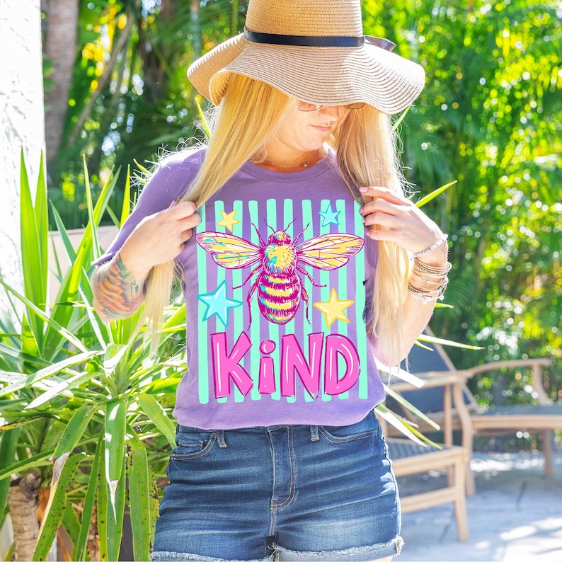 Puede incluir: Una camiseta morada con un gr&aacute;fico de abeja colorida, rayas turquesas y blancas, y la palabra "KIND" en rosa. La abeja tiene alas amarillas, rosas y turquesas. La camiseta se combina con pantalones cortos de mezclilla y un sombrero de paja.