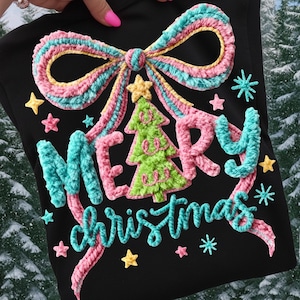 Pode incluir: Camisola preta com um design texturizado e colorido de "Merry Christmas". O design inclui um grande laço, uma árvore de Natal, estrelas e flocos de neve. O texto e os enfeites são rosa, azul, amarelo e verde.